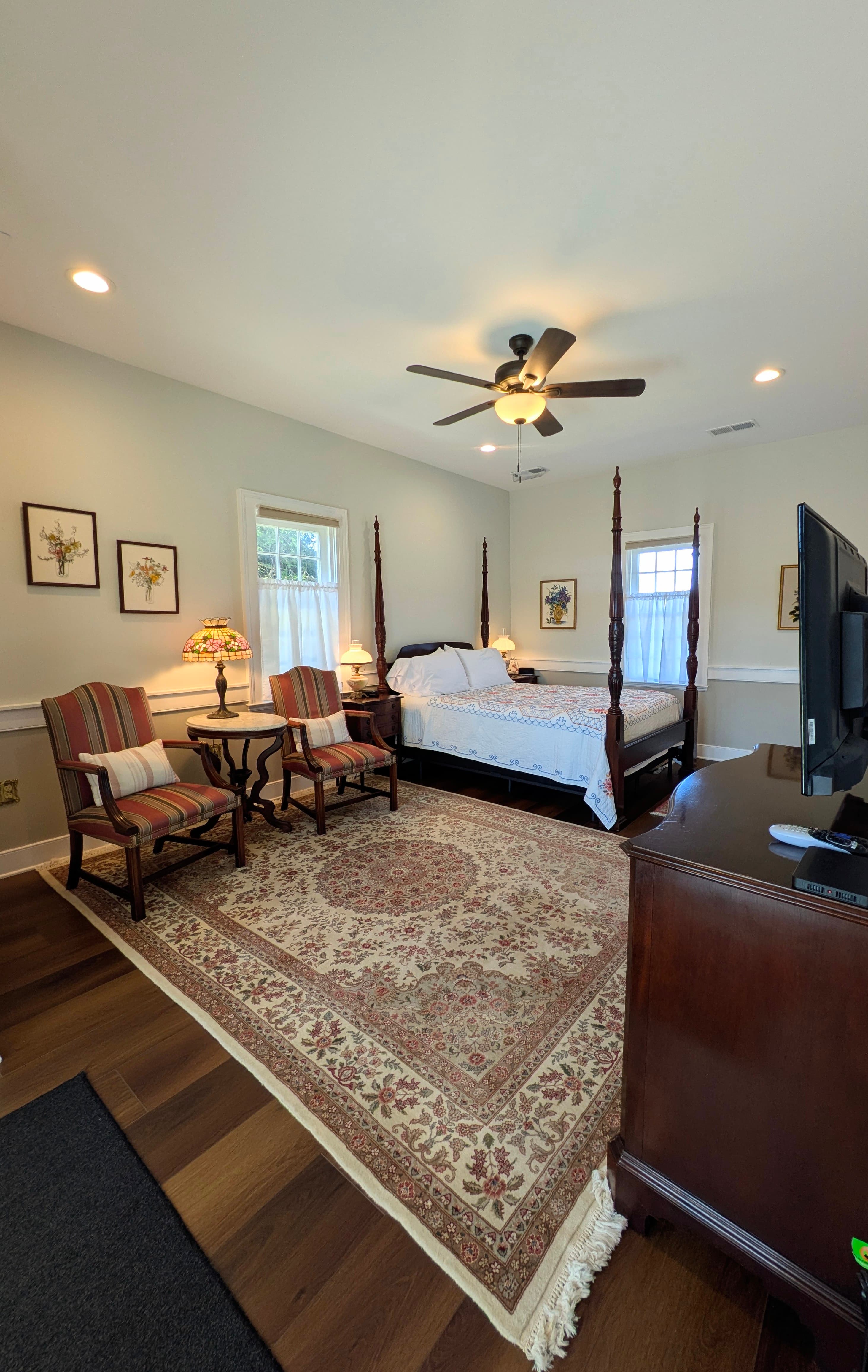 View from doorway: four-poster queen bed, two vintage chairs with vistorian marble-topped side table, wall-mounted TV, dresser, oriental rug