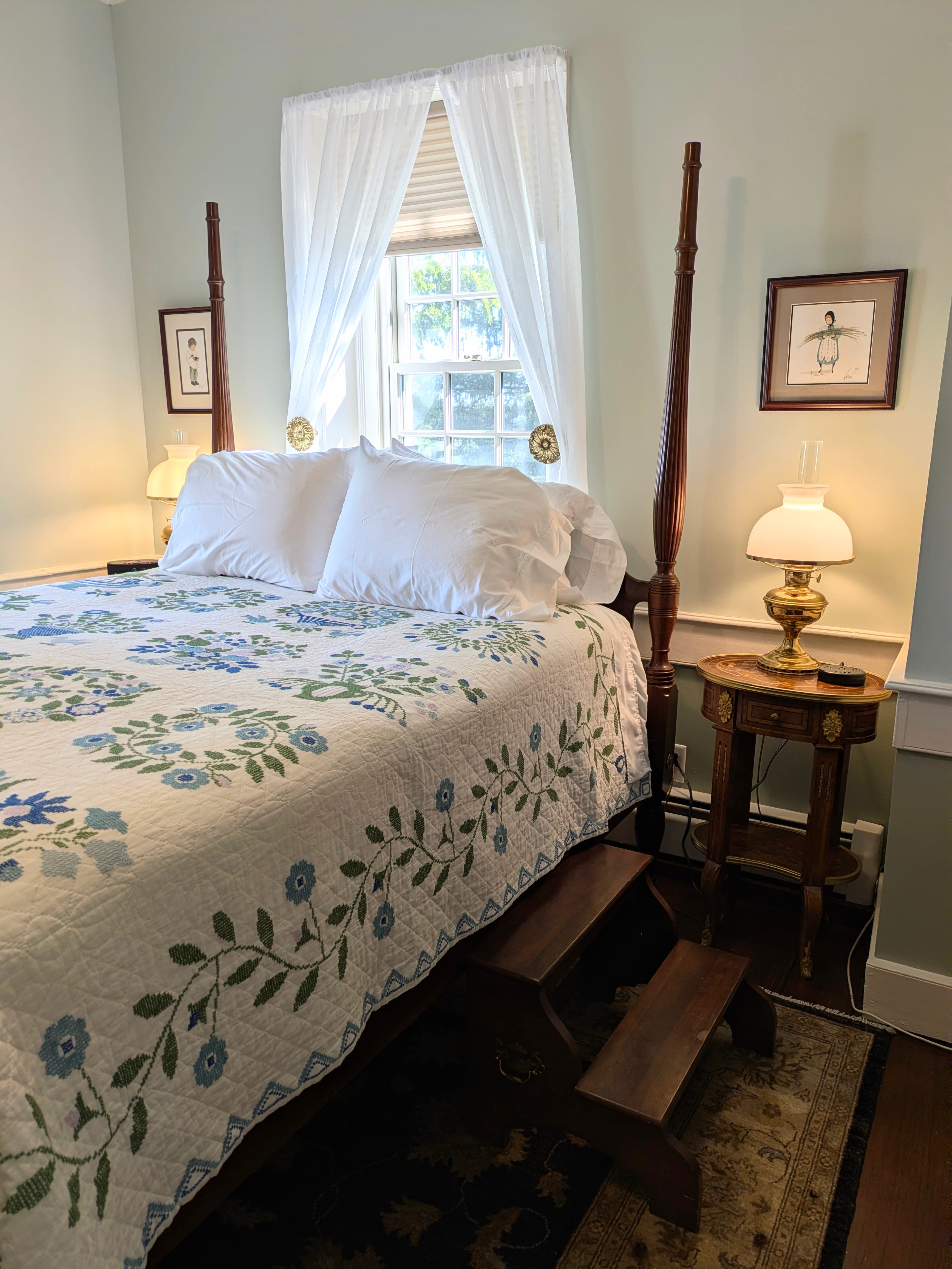 Close up of four poster bed with handmade quilt, window over the bed, two nightstands with lamps