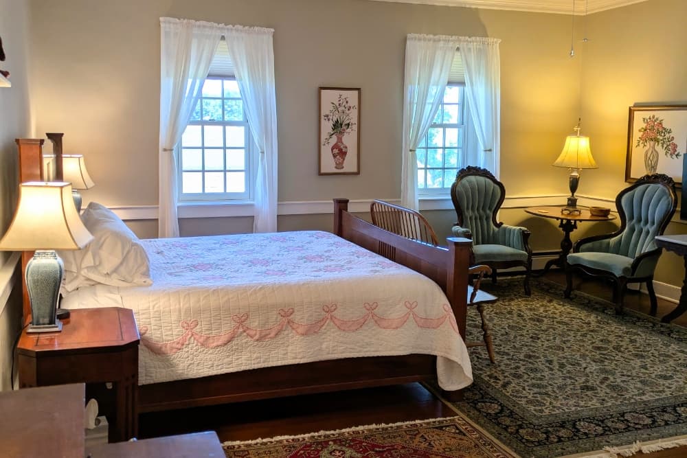 Cozy bedroom with a wooden bed, elegant armchairs, and natural light from two windows.