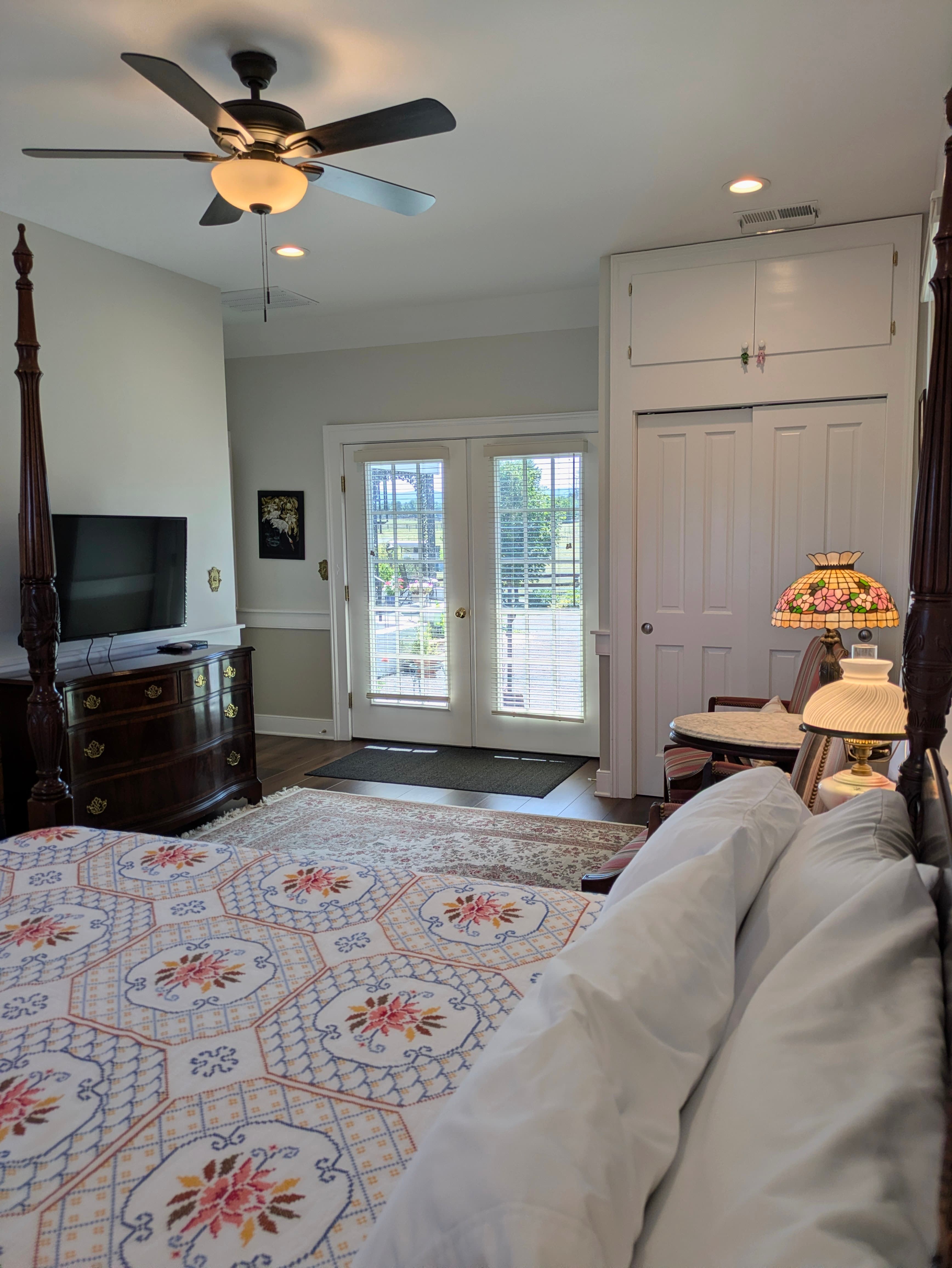 View toward exterior doors with wood-slat venetian blinds, oriental rug, dresser, wall-mounted TV, two vintage guest chairs