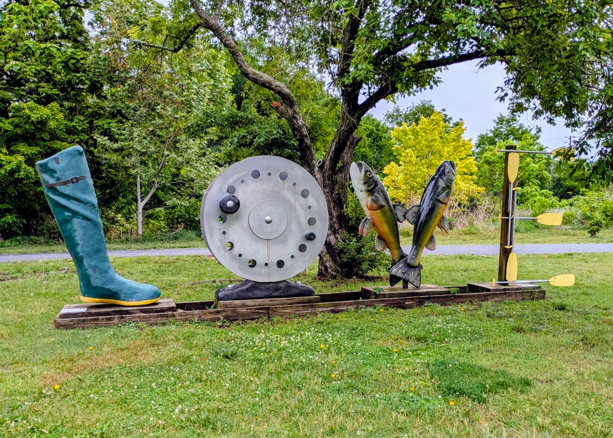 A whimsical outdoor sculpture featuring a large rubber boot, a fishing reel, two fish, and a pair of oars.
