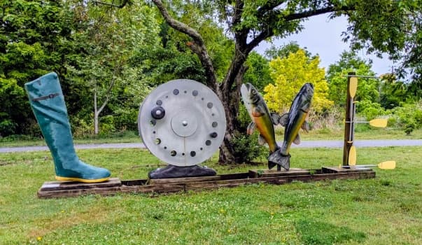 A whimsical outdoor sculpture featuring a giant rubber boot, fishing reel, two fish, and a paddle, set against a lush green background.