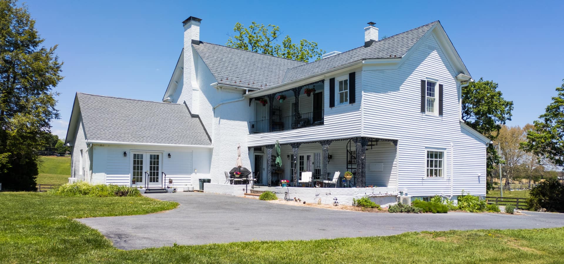 A white two-story farmhouse with a large porch, surrounded by green grass and trees under a clear blue sky.