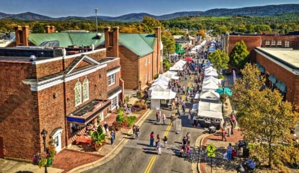A bustling street festival with vendors beneath white tents, surrounded by mountain views.