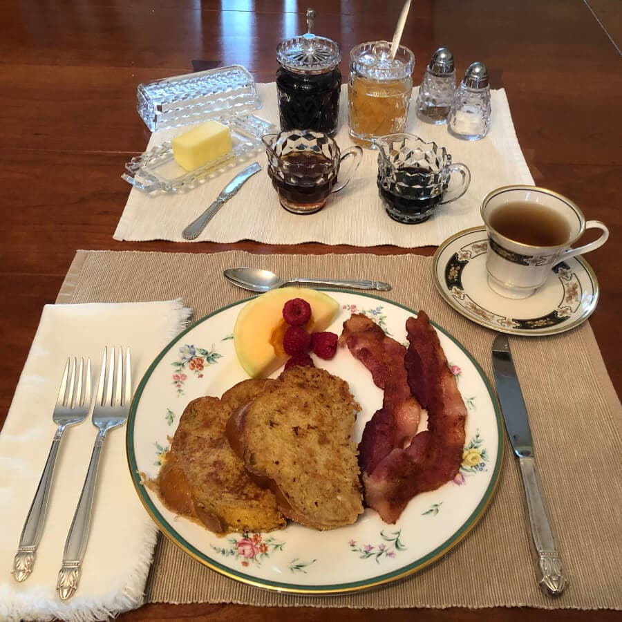 A plate with French toast, bacon, and fresh fruit beside a cup of coffee and various condiments on the table.