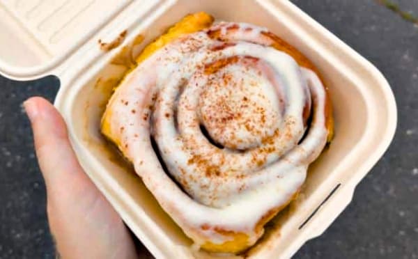 A hand holds a cinnamon roll with icing in a takeout container.