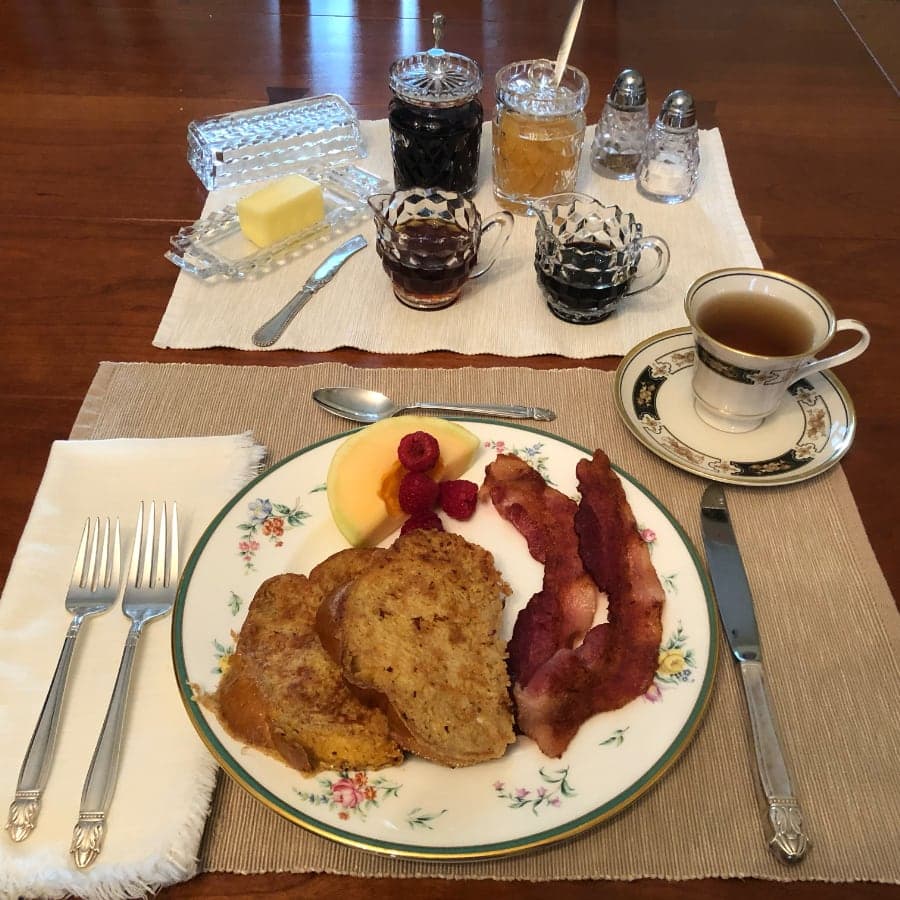 A beautifully arranged breakfast plate featuring French toast, bacon, fruit, butter, assorted jams, and a cup of tea on an elegant table setting.