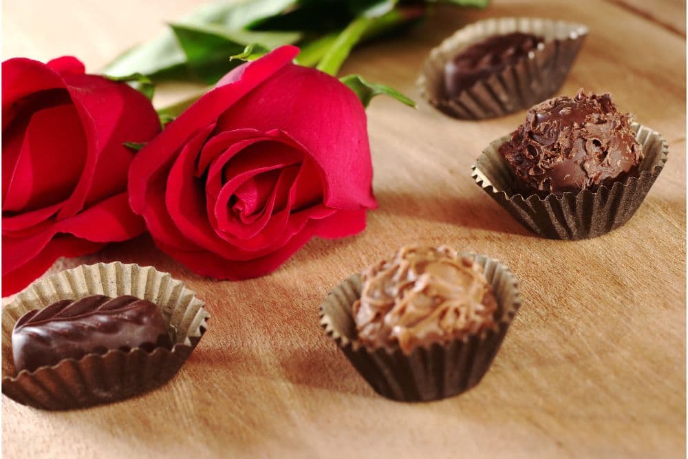 A pair of red roses beside various chocolate truffles on a wooden surface.