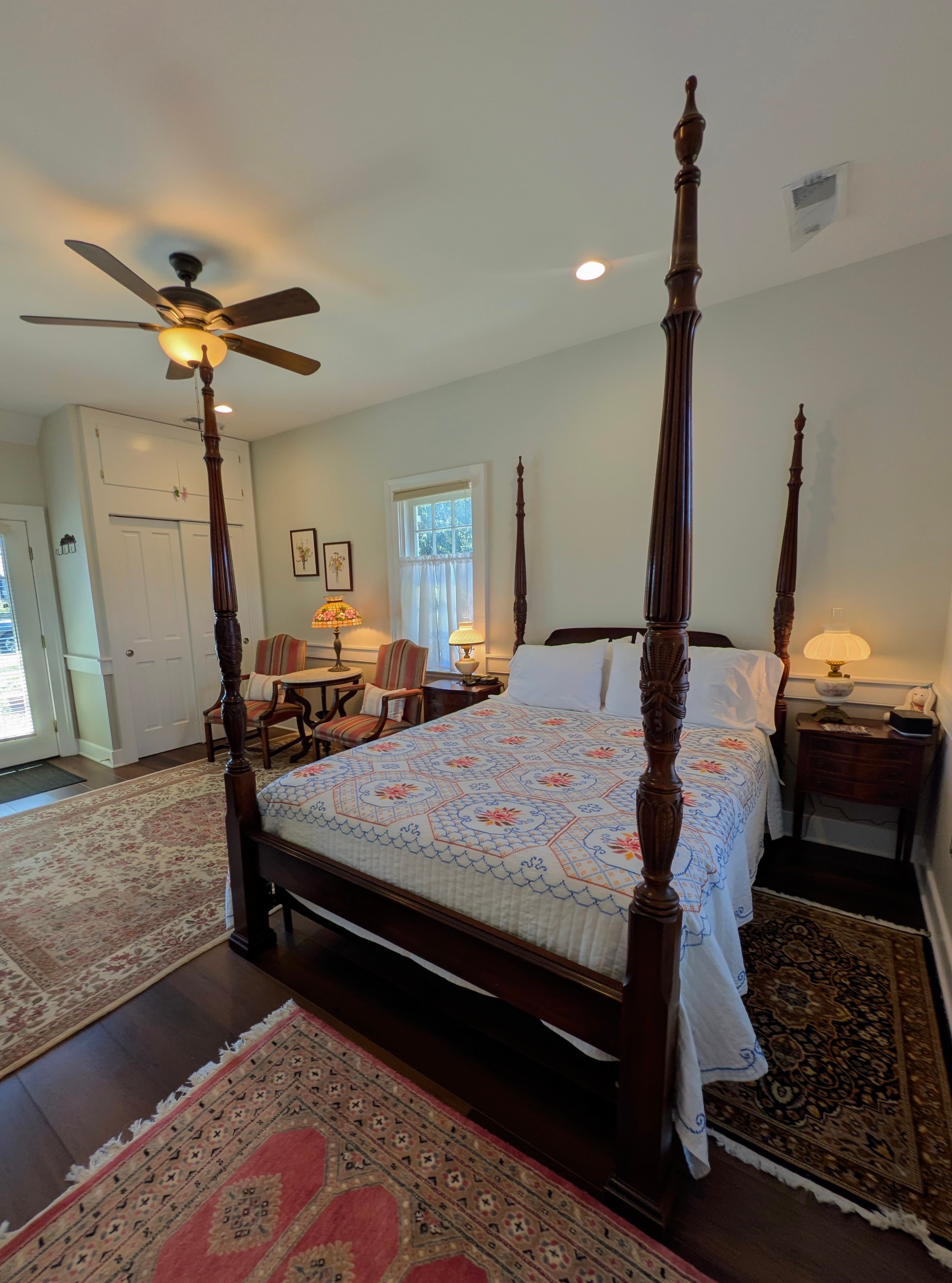 Four poster bed, two vintage guest chairs with victorian marble-top end table, oriental rug, two windows.