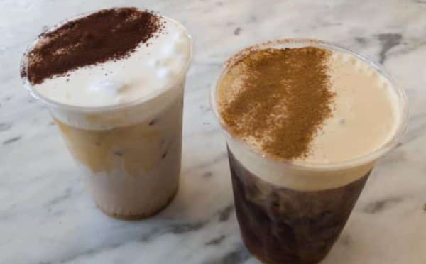 Two cups of iced coffee topped with different cocoa powders on a marble surface.