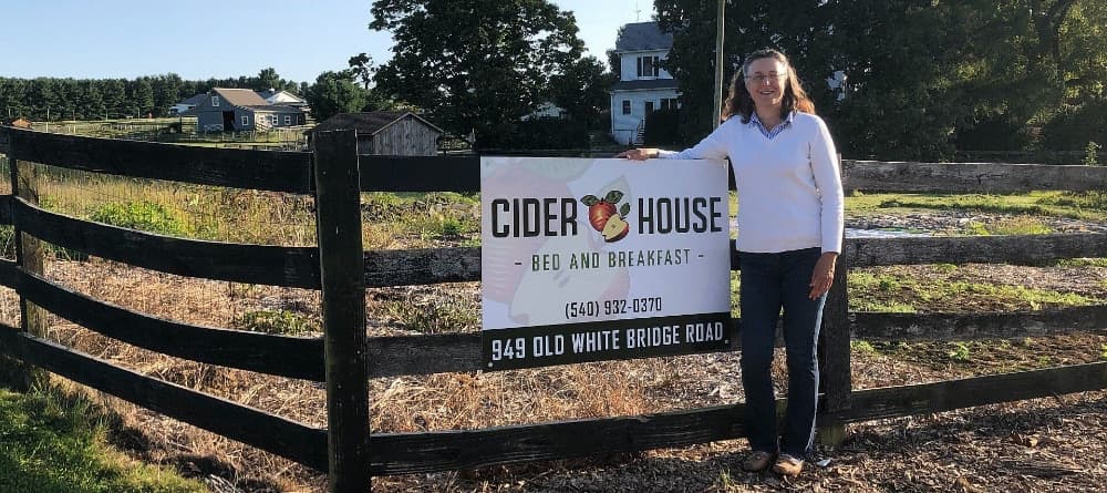 A woman stands beside a sign for "Cider House Bed and Breakfast" near a rustic fence in a rural setting.