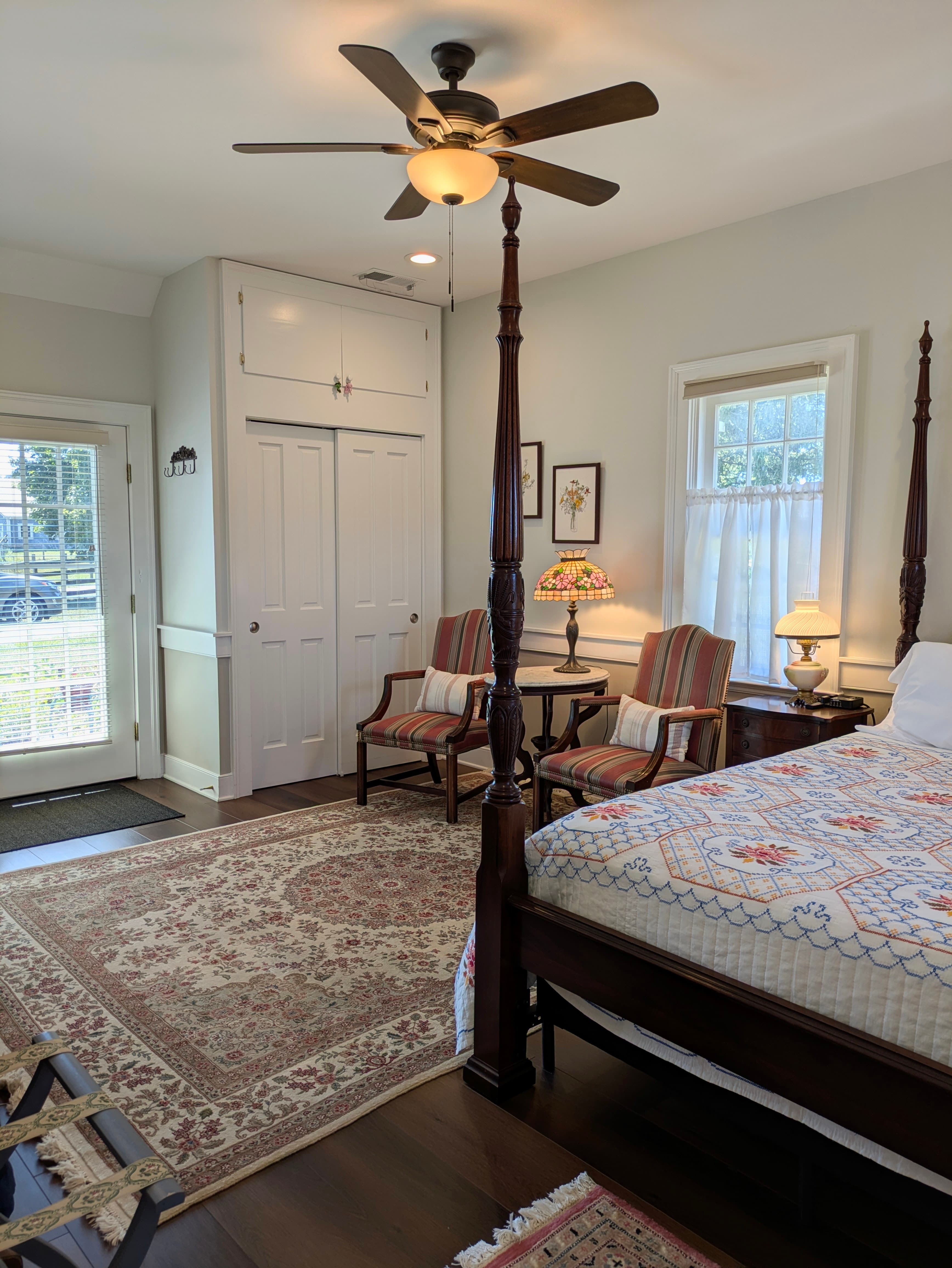 Exterior door, closet, coat hooks, sitting area with 2 chairs and endtable, window, bed with homemade quilt