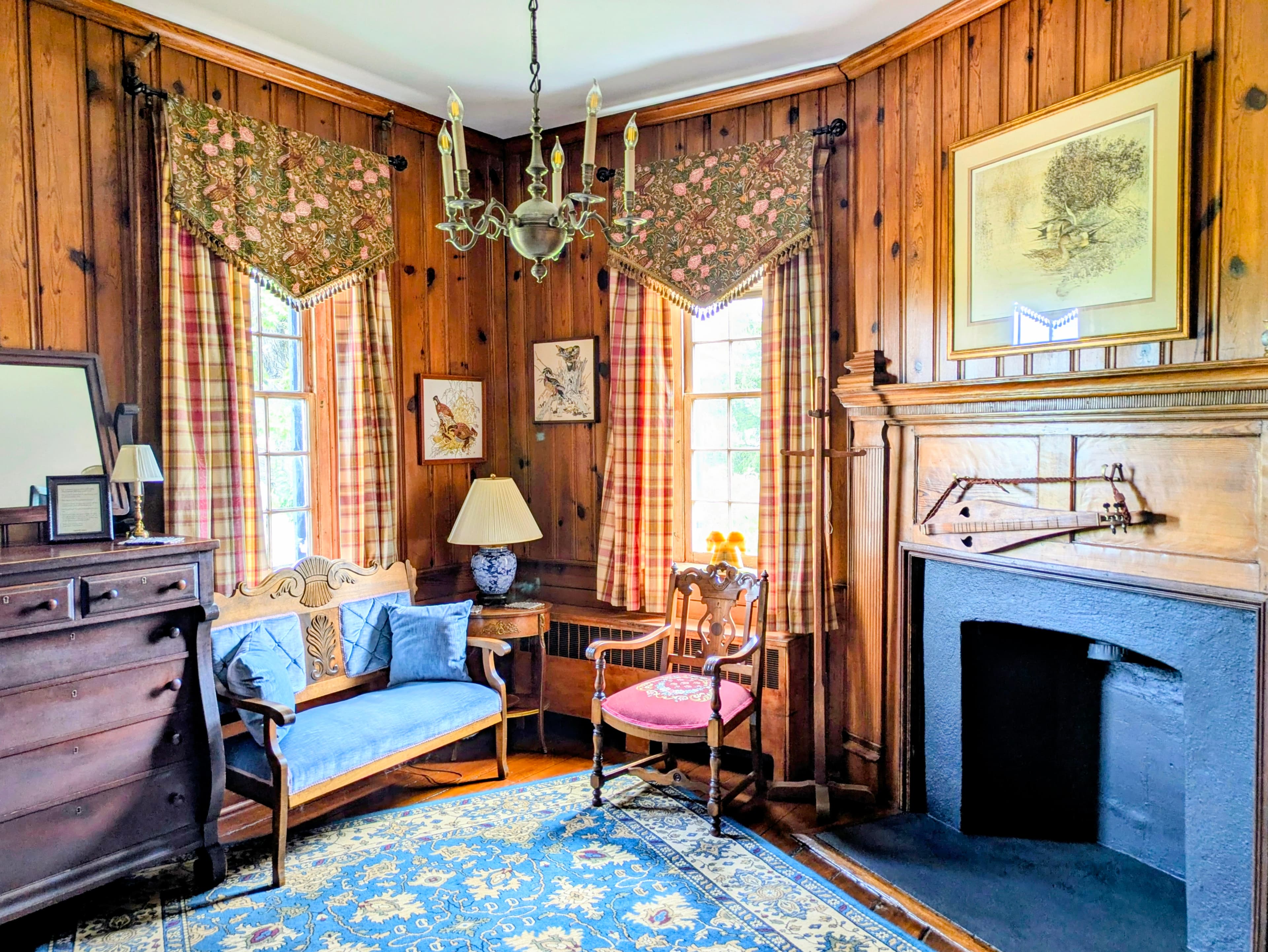 Entry sitting room of suite with wood panelling, dresser, tall windows with drapers, fireplace with mantel and picture of ducks over the mantel