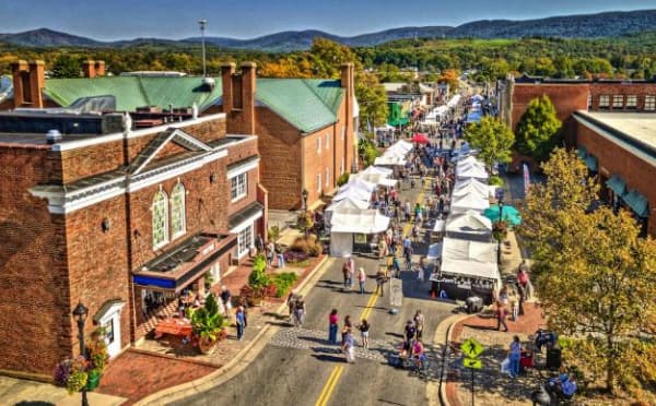 Aerial view of a bustling street fair with vendor tents, people walking, and autumn foliage in the background.