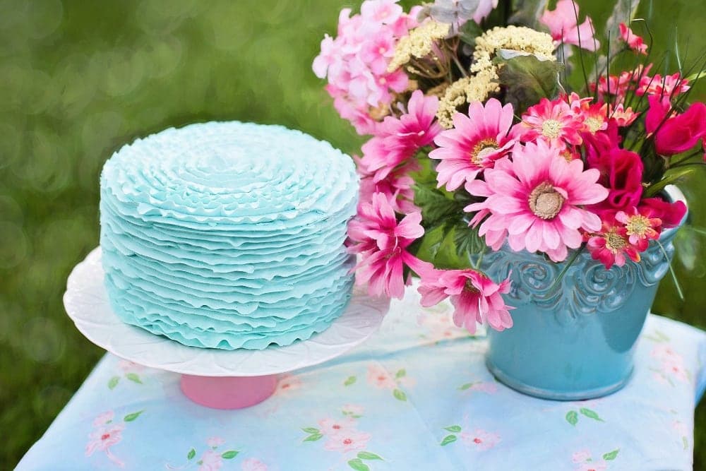 A blue ruffled cake sits on a white plate next to a vase of vibrant pink flowers.