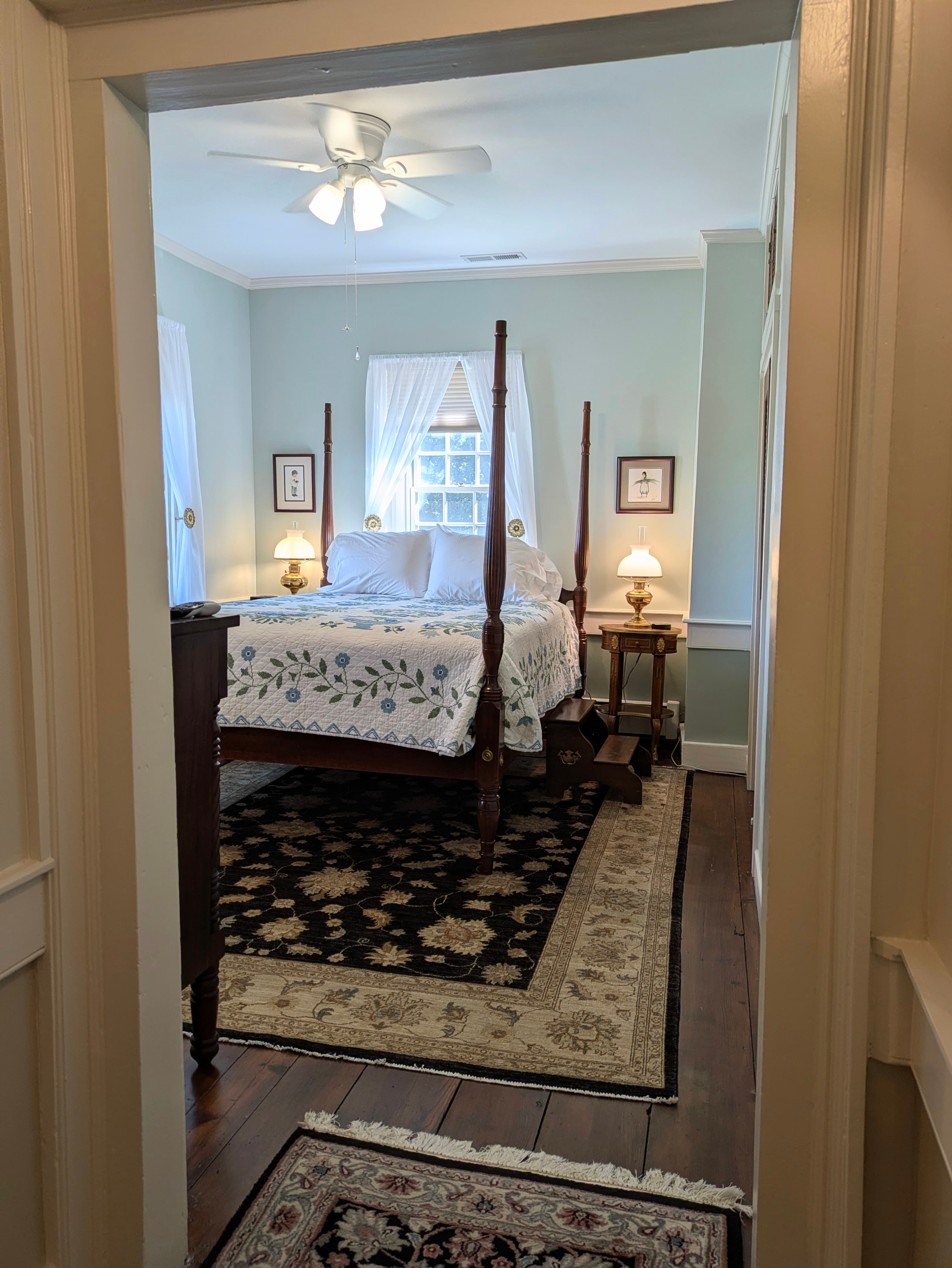 Viewing the room through the hall doorway: queen four-poster bed, window above bed, nightstands with lamps, oriental rug
