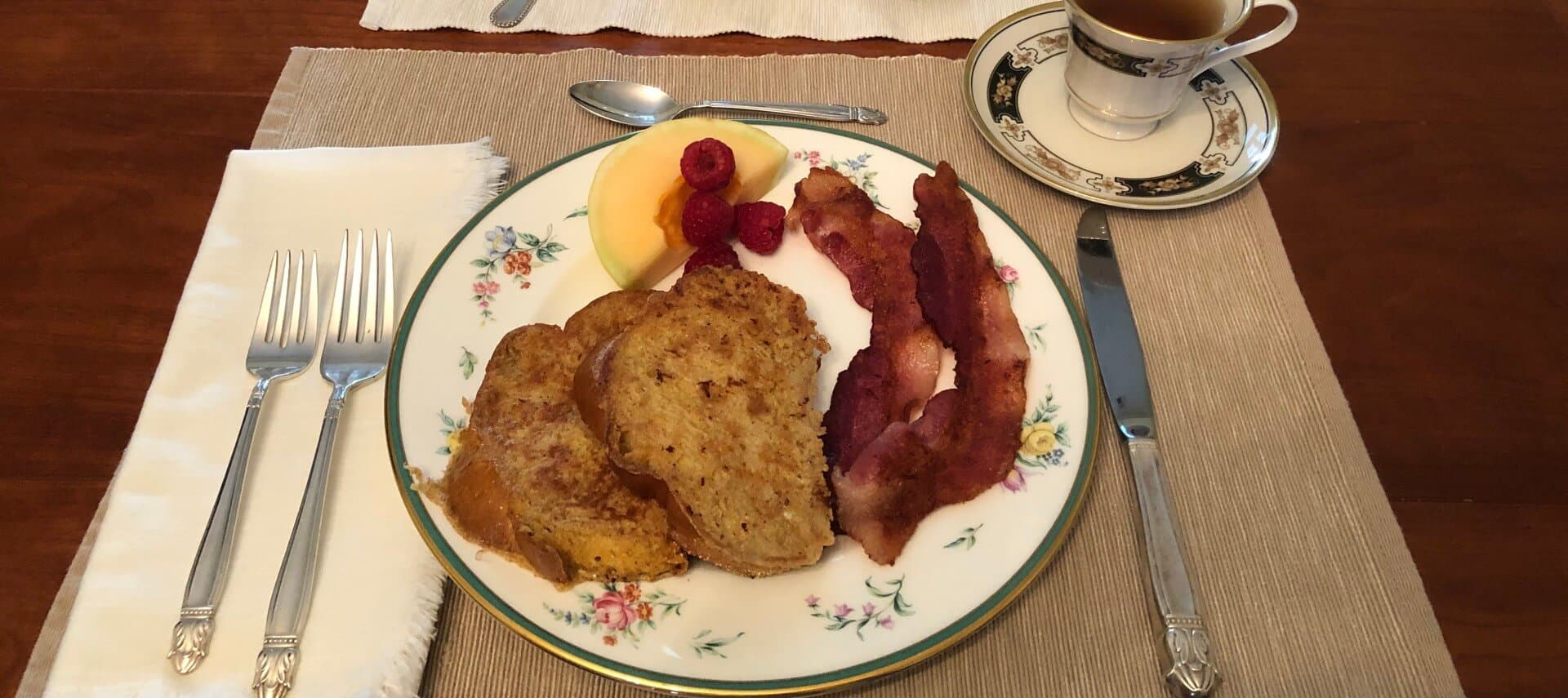 A plate with French toast, bacon, fresh fruit, and a cup of coffee set on a table.