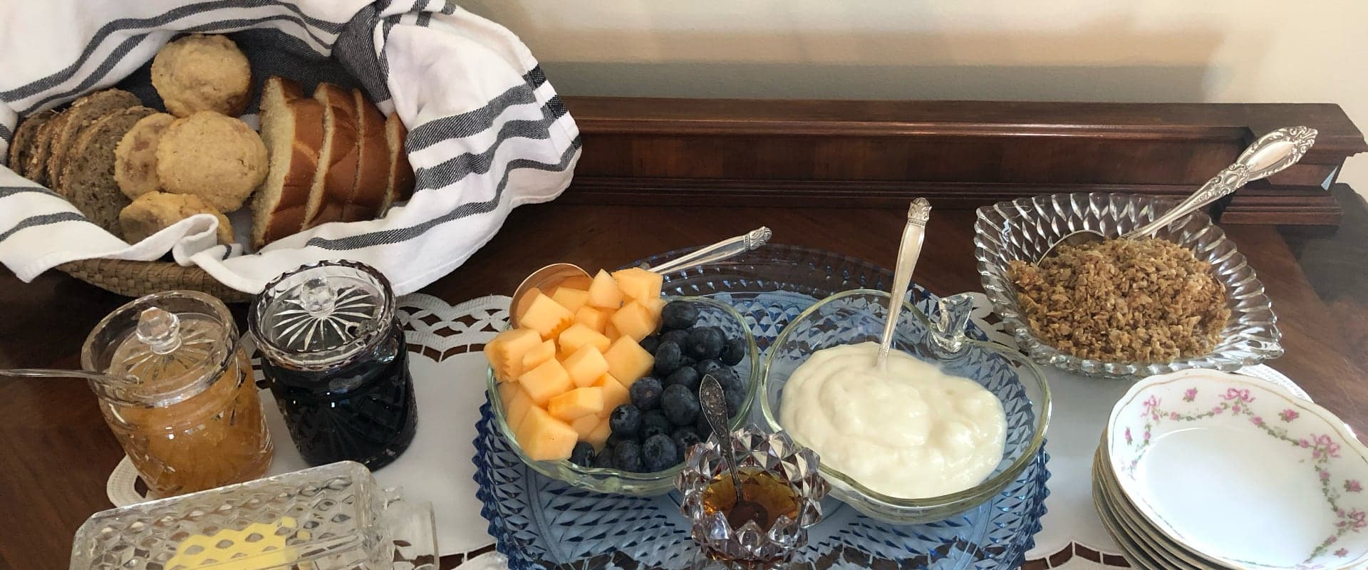 A breakfast spread featuring cookies, sliced bread, fruit, yogurt, granola, and jars of jam on a lace tablecloth.