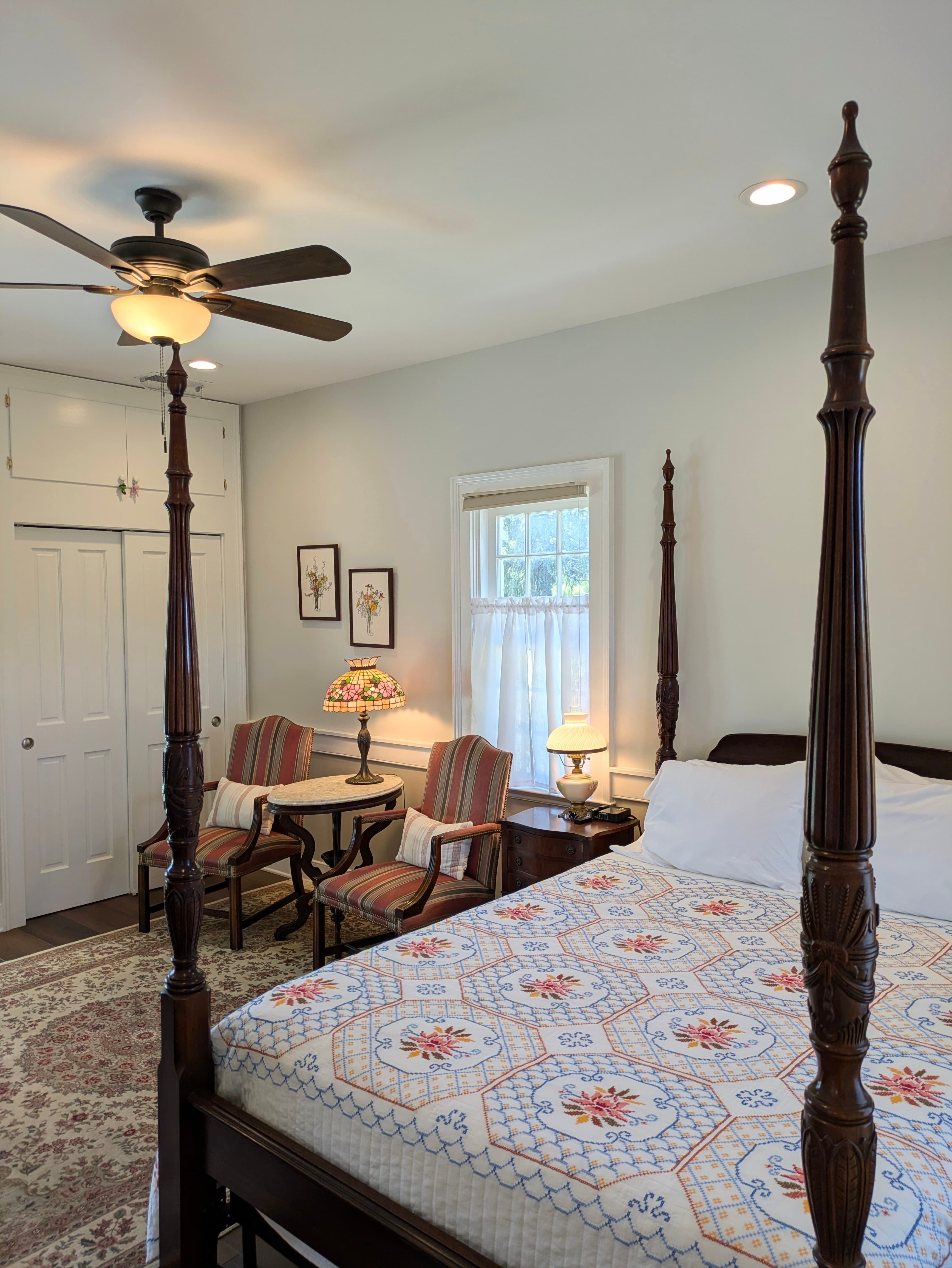 View toward window with four-poster bed, 2 vintage chairs, marble-topped side table, tiffany-style lamp, closet