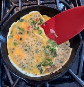 A red spatula lifts a partially cooked omelet with vegetables in a cast iron skillet on a stove.