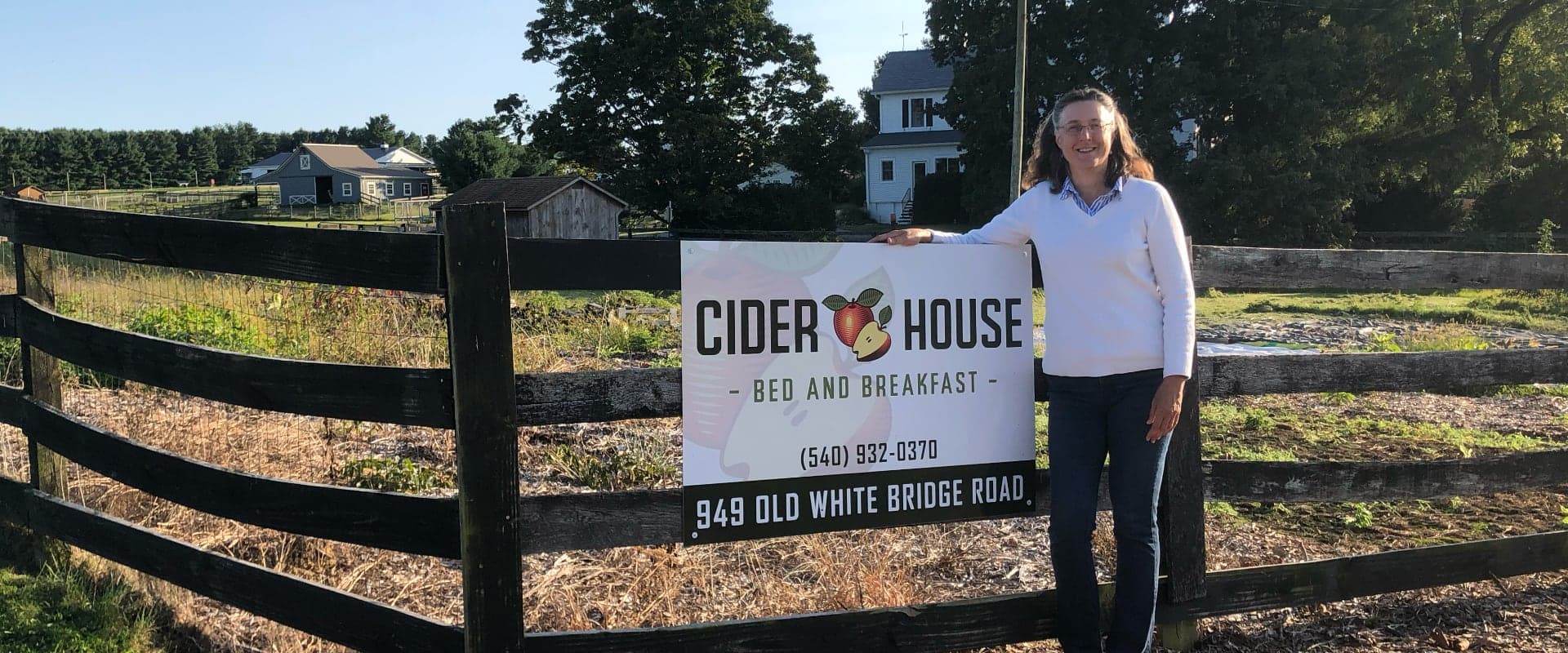 A woman stands beside a sign for "Cider House Bed and Breakfast" on a farm.