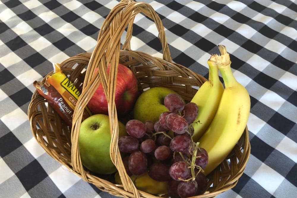 A woven basket filled with an apple, green apples, bananas, grapes, and a snack bar, resting on a checkered tablecloth.