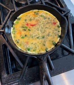 A colorful vegetable omelette cooking in a cast iron skillet on a stovetop.
