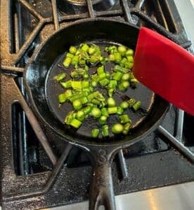 Chopped green vegetables are sautéing in a cast iron skillet with a red spatula.