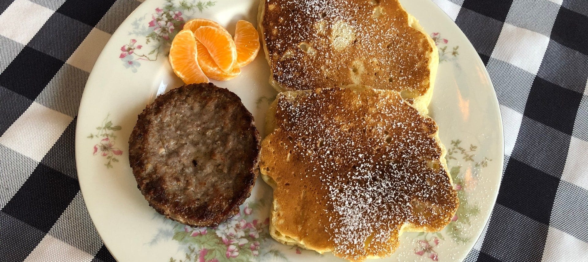 Two apple pancakes sprinkled with powedered sugar, a sausage patty, and some orange sections on a plate.