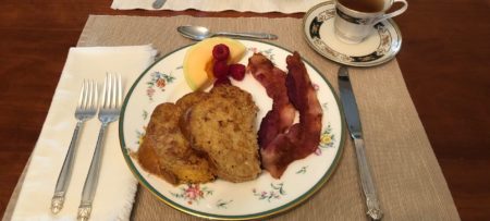Two slices of french toast, two slices of bacon, and some cantaloupe and berries on fine china on a placemat with a sliver place setting.