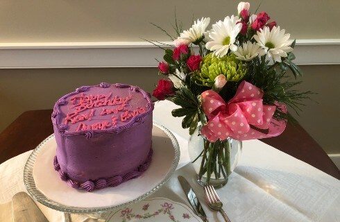 Purple birthday cake on a pedestal and a bouquet of flowers in a vase.