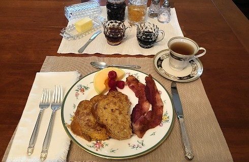 Table setting for one with plate holding French toast, bacon and fruit with syrup, jams, and butter