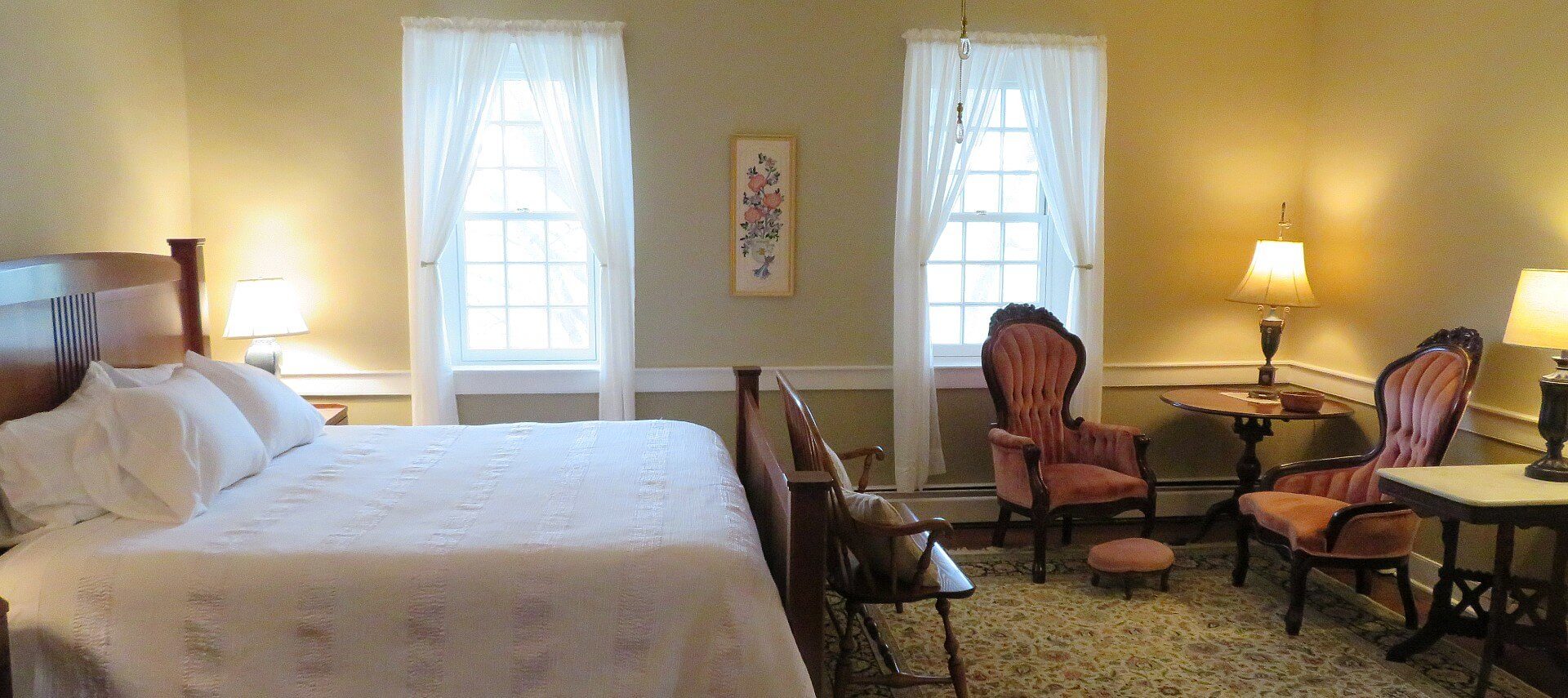Guest room with king bed in white linens, sitting area and two bright large windows with white curtains