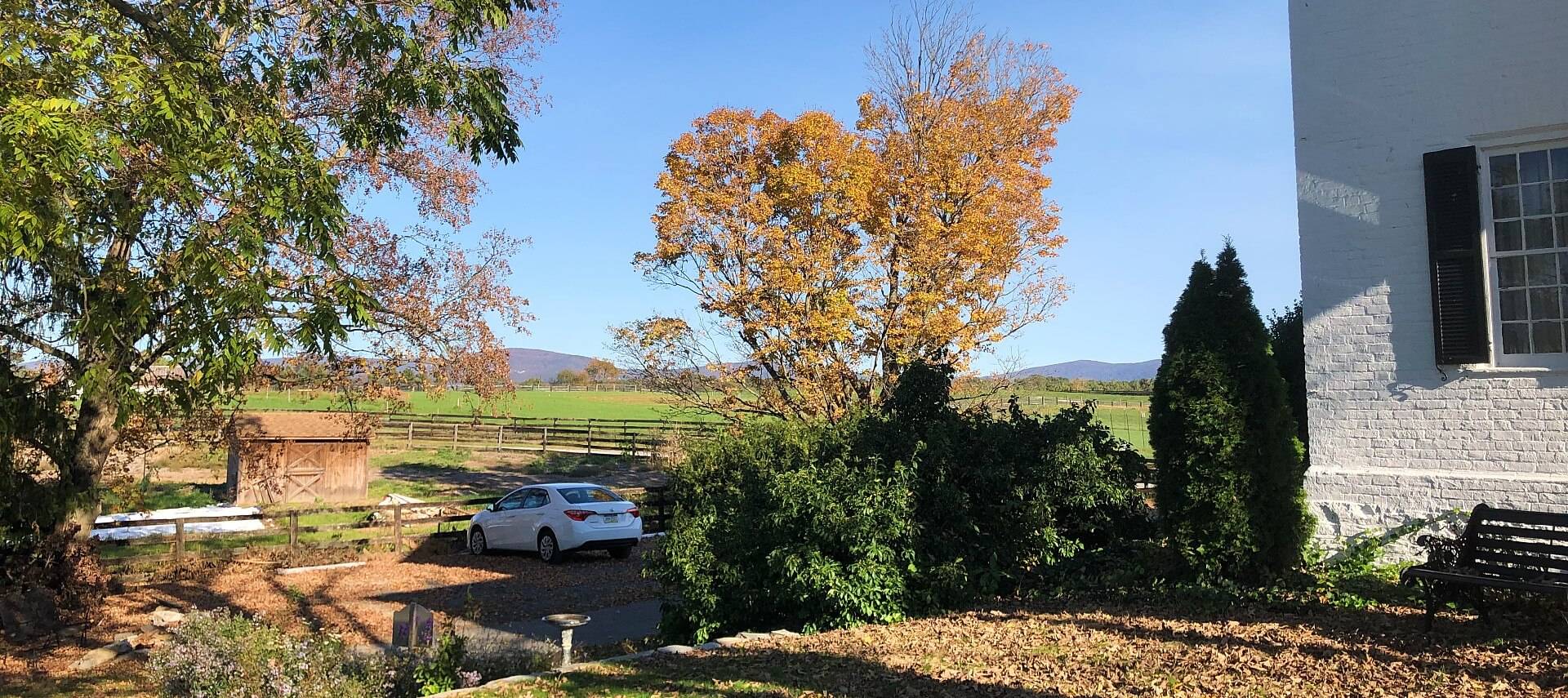 Side yard of a white brick home with fenced farmland and a parked white car